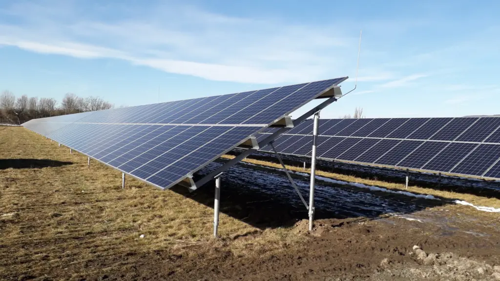 A ground-level shot of a large solar panel array on a grassy field under a bright, blue sky with some scattered clouds. The panels are tilted up on silver metal frames. The ground is dry and patchy with some areas of dirt and a few patches of snow, suggesting it is a cold or dry season.