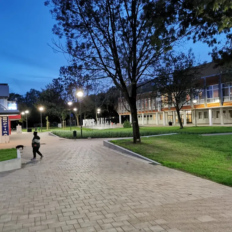 The image shows an evening or early night scene in an outdoor urban area with paved walkways, grass patches, and trees. Streetlights are illuminated, casting warm light around the area. A person dressed in dark clothing is seen walking with a cat nearby. On the left, there is a small building or kiosk with signage including "OPTIKA" and some advertising posters in the windows. In the background, a larger building with many windows is visible, with some interior lights on. A water fountain with jets of water is also visible near the center, adding to the ambiance of the scene. The sky is a deepening blue, indicating dusk.