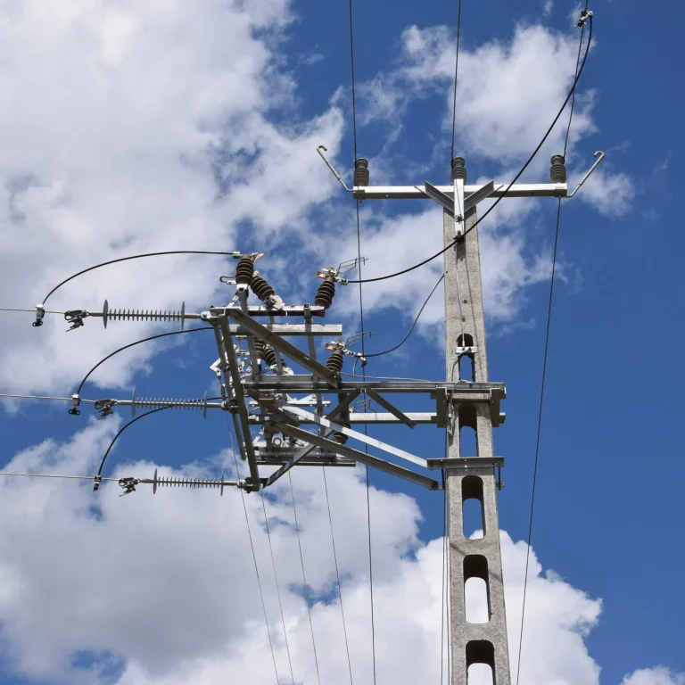 A low-angle shot of a gray concrete electrical utility pole against a bright blue sky with scattered clouds. The pole supports a complex arrangement of wires, insulators, and metal crossarms, which are silhouetted against the sky. A bundle of thick, insulated wires runs horizontally across the top, while other wires and insulators extend diagonally from the pole.